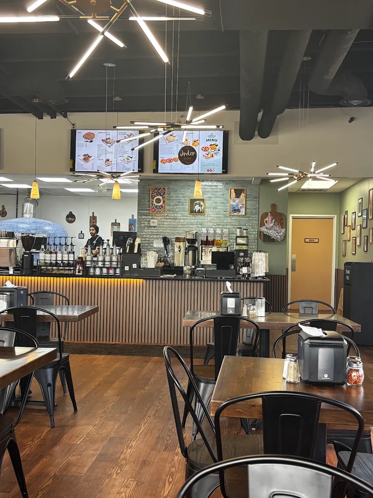 The finished Al Manakeesh dining room in Fishers: a blue-tiled wood-fired oven behind the slatted service counter, linear pendant fixtures over the dining floor, and backlit menu boards above the order point.